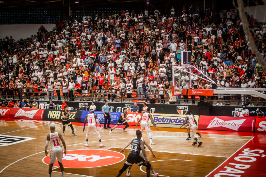 Foto de uma quadra de basquete, onde ocorre um jogo de basquete, em quadra estão 8 jogadores. Ao fundo centenas de torcedores acompanham o jogo em uma das arquibancadas localizadas acima do nível da quadra.